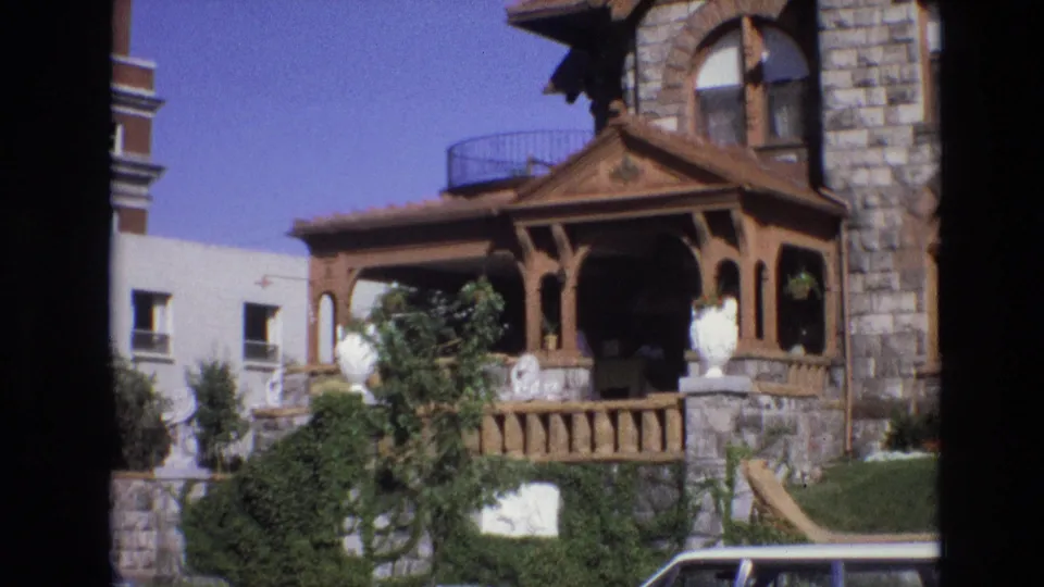 1974 Denver home-movie frame centered on the house porch and front wall, with carved wood trim, ivy-covered stonework, clipped grass, a neighboring building, and a partial parked car visible.