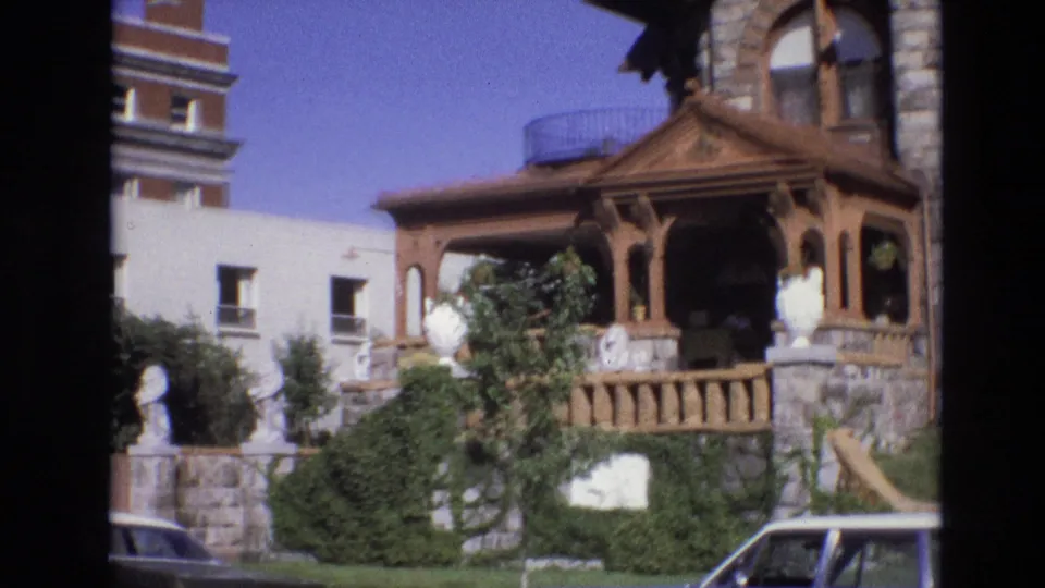 1974 Denver exterior frame showing the ornate porch from farther along the pan, with shrubs climbing the stone frontage, white decorative urns or statues, blue sky, and part of a car at the lower right.