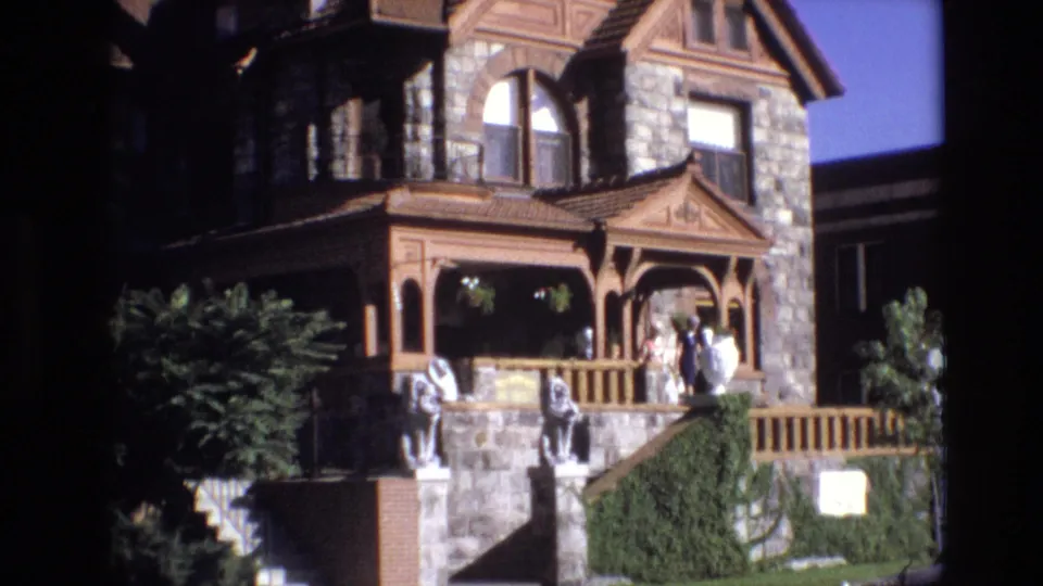 1974 Denver home-movie still with a closer look at the ornate porch, stone facade, arched upper window, people by the entrance, steps, statues, and surrounding plants.