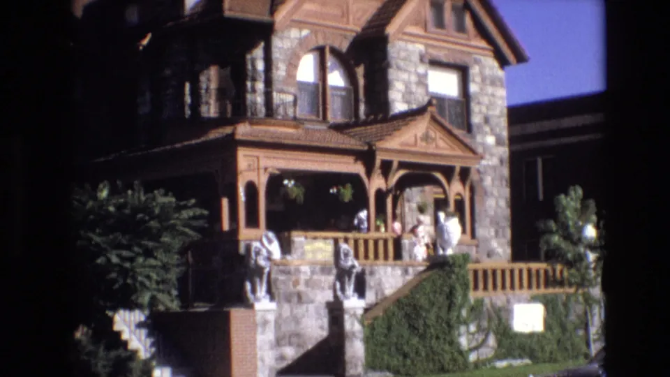 1974 Denver frame showing the stone-and-wood house exterior, porch visitors, decorative steps with statues, green shrubs, and a bright blue sky.