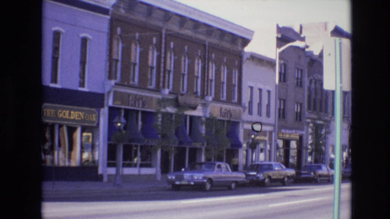 1974 Denver downtown street view with commercial storefronts, blue awnings, The Golden Oak sign, and several parked cars along the curb.