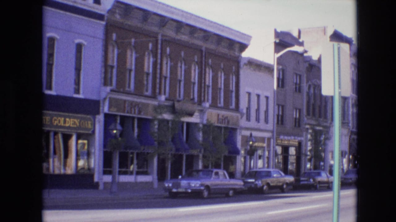 1974 Denver street scene showing a row of downtown shops with visible signage, blue awnings, parked sedans, and a pickup truck in bright daylight.