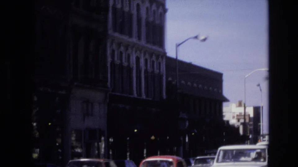 Later frame from the 1974 Denver street sequence showing a darker view of a tall commercial facade, parked cars, a pickup truck, and streetlights under bright sky.