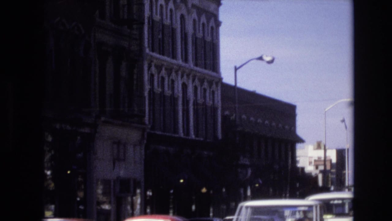 Denver street view from 1974 metadata showing passing vehicles, streetlights, and ornate multi-story commercial buildings in soft 8mm color.