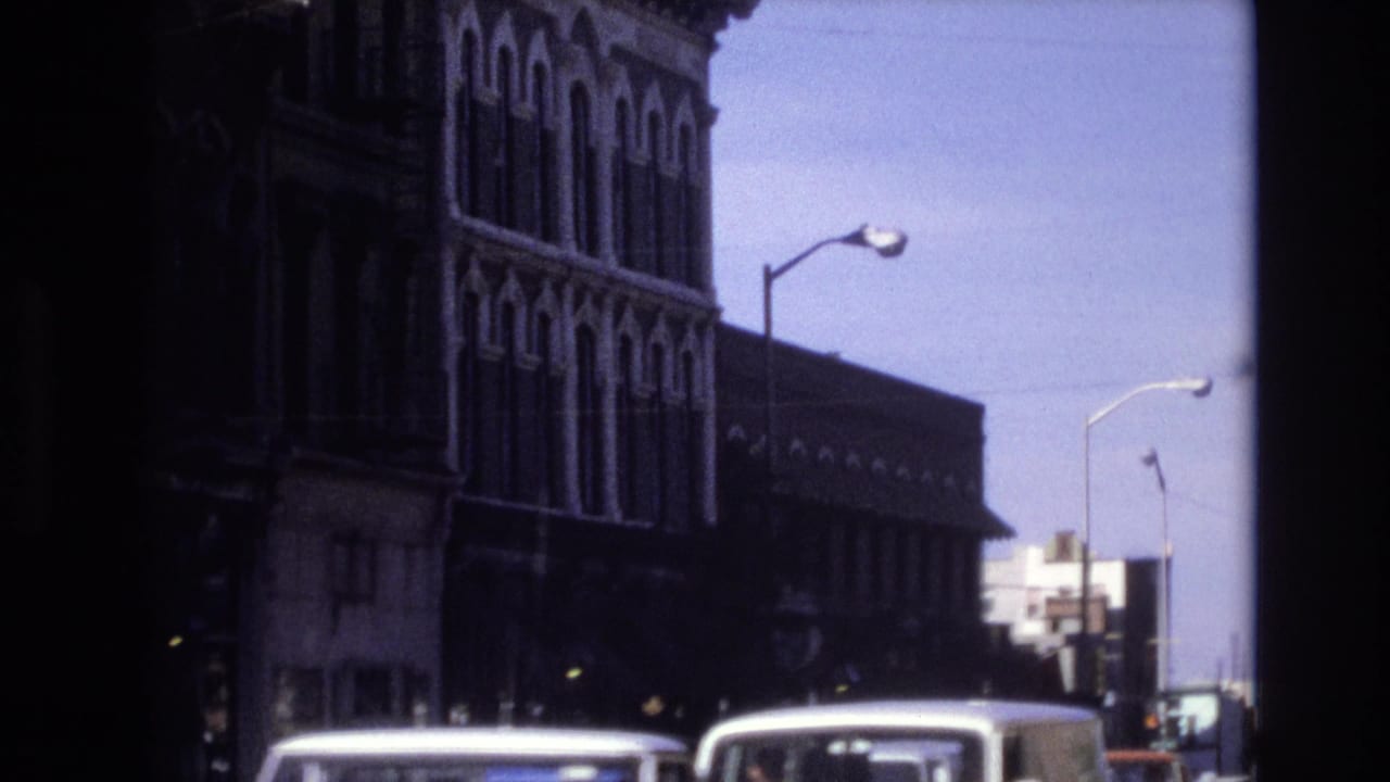 Vehicles move along a street beside tall masonry buildings and streetlights.