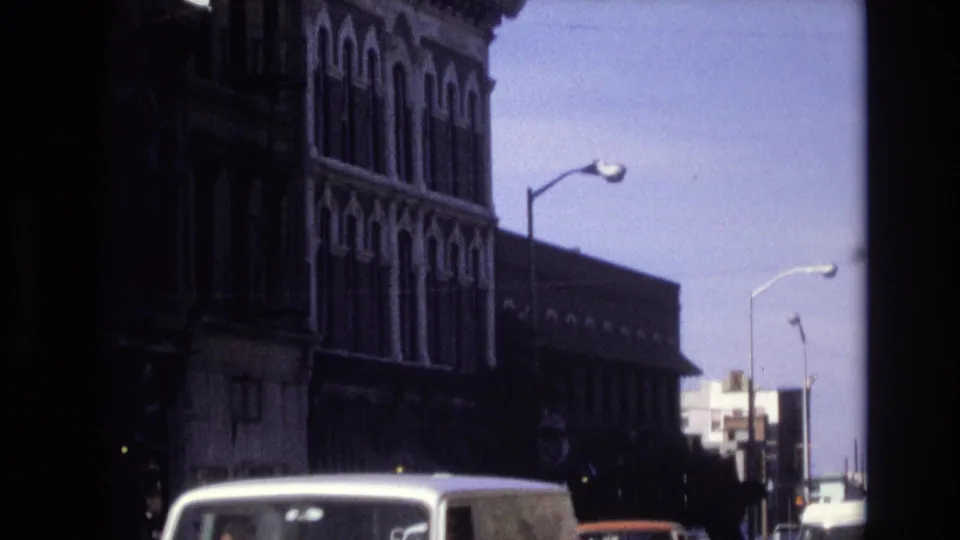Denver 1974 home-movie frame with a white van crossing the lower foreground and older commercial architecture rising behind it.