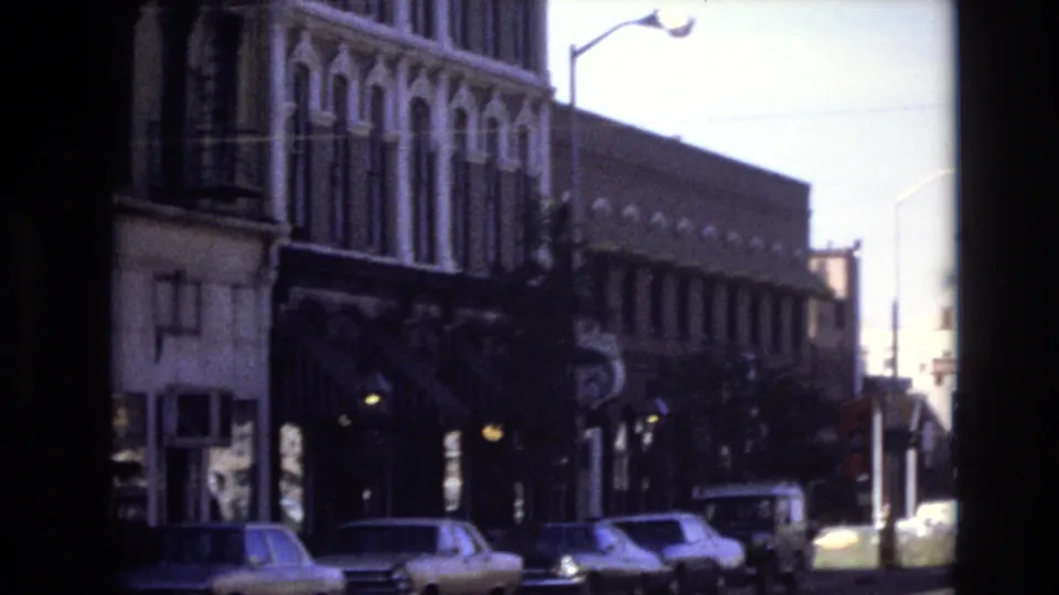 1974 Denver street view with cars near the curb, a streetlight, and ornate older building fronts captured in grainy 8mm color.