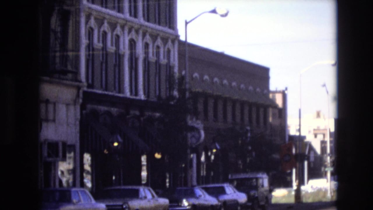 1974 Denver home-movie frame showing parked cars along a curb beneath older storefront facades with ornamental details and a streetlight.