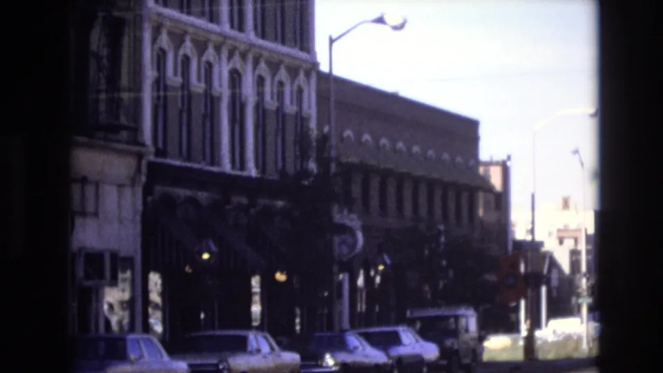 1974 Denver home-movie frame showing parked cars along a curb beneath older storefront facades with ornamental details and a streetlight.