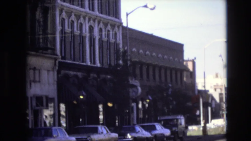 1974 Denver 8mm frame showing a quiet downtown block with parked cars, older commercial facades, a streetlight, and deep storefront shadows.
