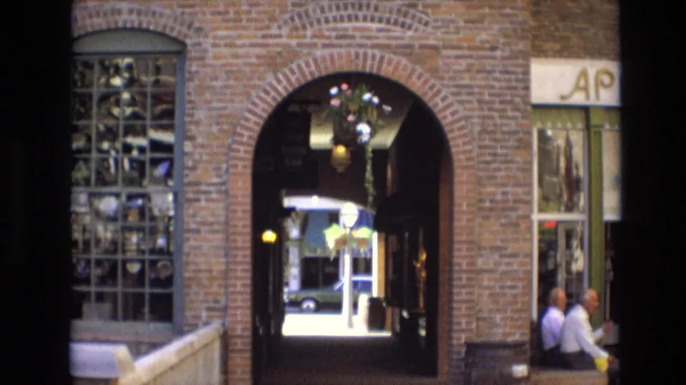 1974 Denver frame showing a brick commercial archway, glass-block window, hanging flower basket, storefront signage fragment, and two men seated near the entrance.