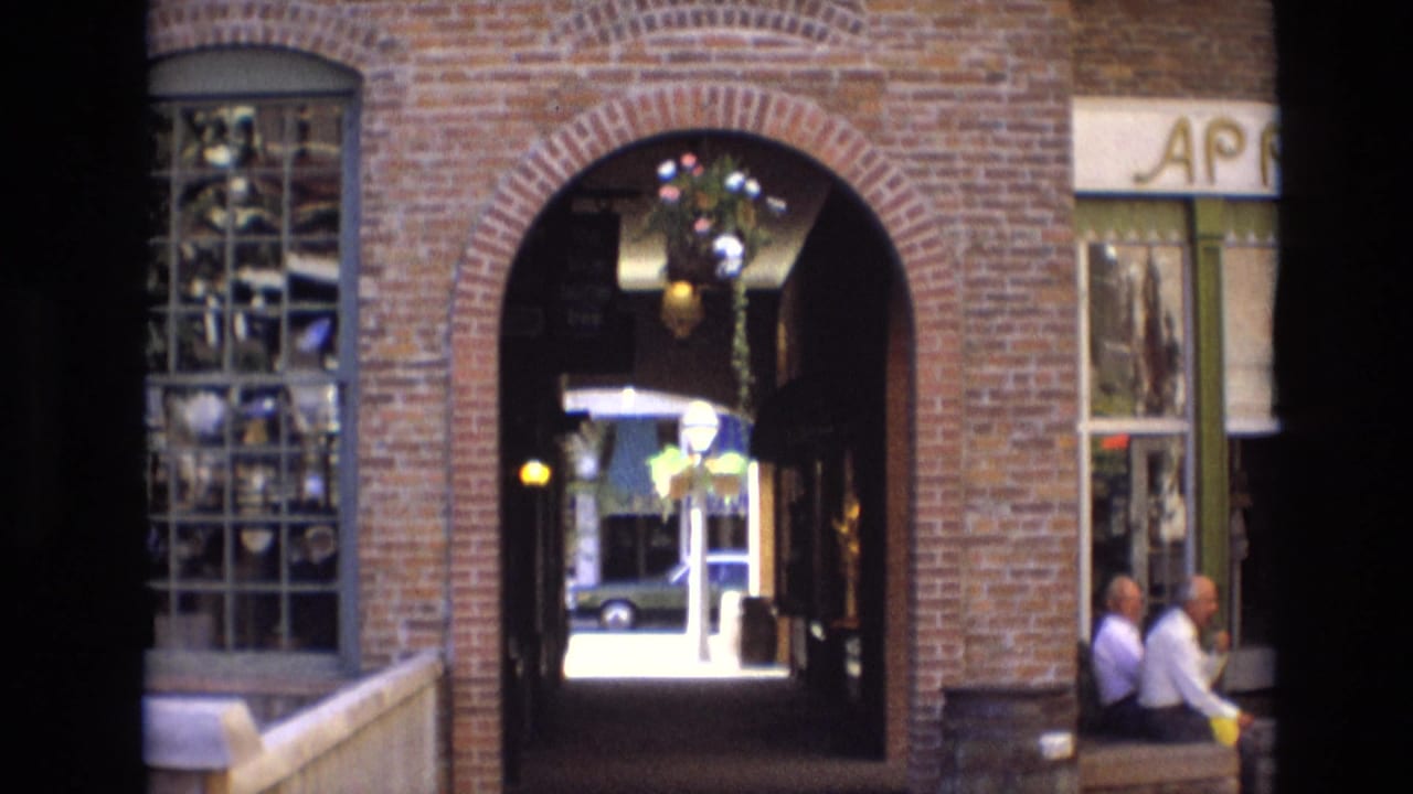 1974 Denver home-movie frame showing a brick Larimer Square archway, display window, covered passage, and two older men seated near the storefront edge.