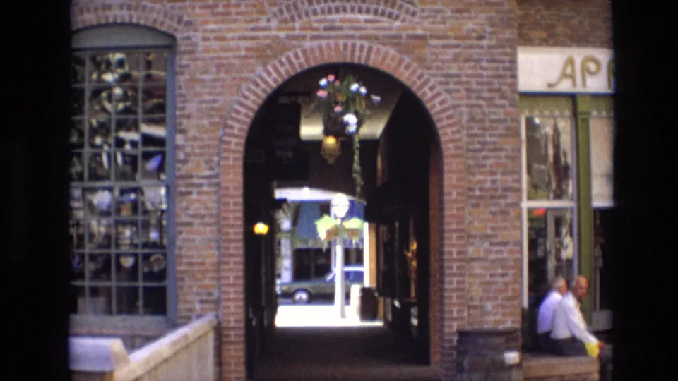 1974 Denver footage frame with a brick arch into Larimer Square, reflective shop window, hanging basket, and two men seated beside partial storefront lettering.