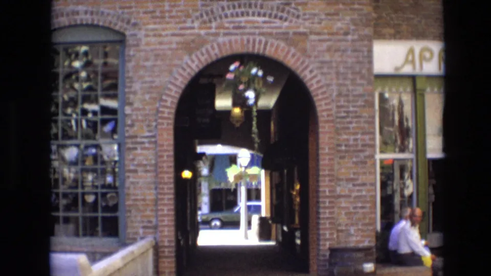 1974 Denver home-movie view of the brick Larimer Square passage, shop display window, hanging flowers, and two older men seated in conversation near the storefront.