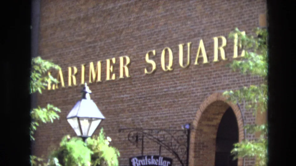 1974 Denver frame confirming Larimer Square with gold lettering on a brick facade, a white street lamp, foliage, and an arched doorway below.