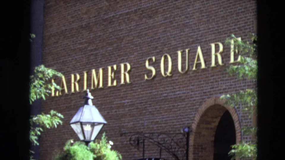 1974 Denver 8mm still focused on the Larimer Square brick facade with gold lettering, leafy branches, a foreground lamp, and an arched doorway.