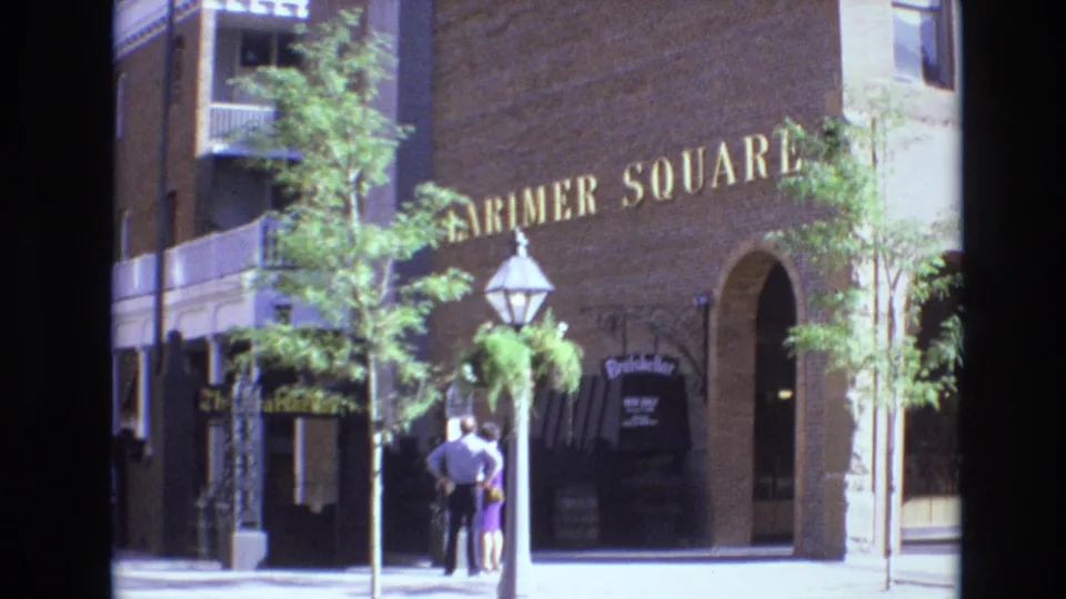 Denver 1974 home-movie frame looking toward Larimer Square, with the brick building, gold lettering, lantern post, trees, hanging business sign, and two adults on the sidewalk.