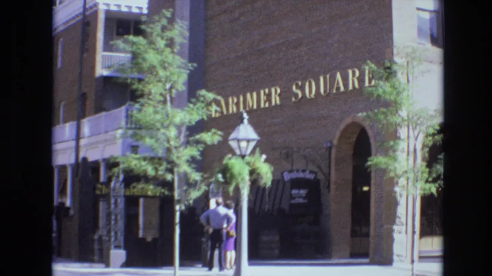 Denver 1974 frame showing the Larimer Square brick facade with gold wall lettering, a lantern post, young trees, a Brauhaus-style hanging sign, and two adults standing near the entrance.