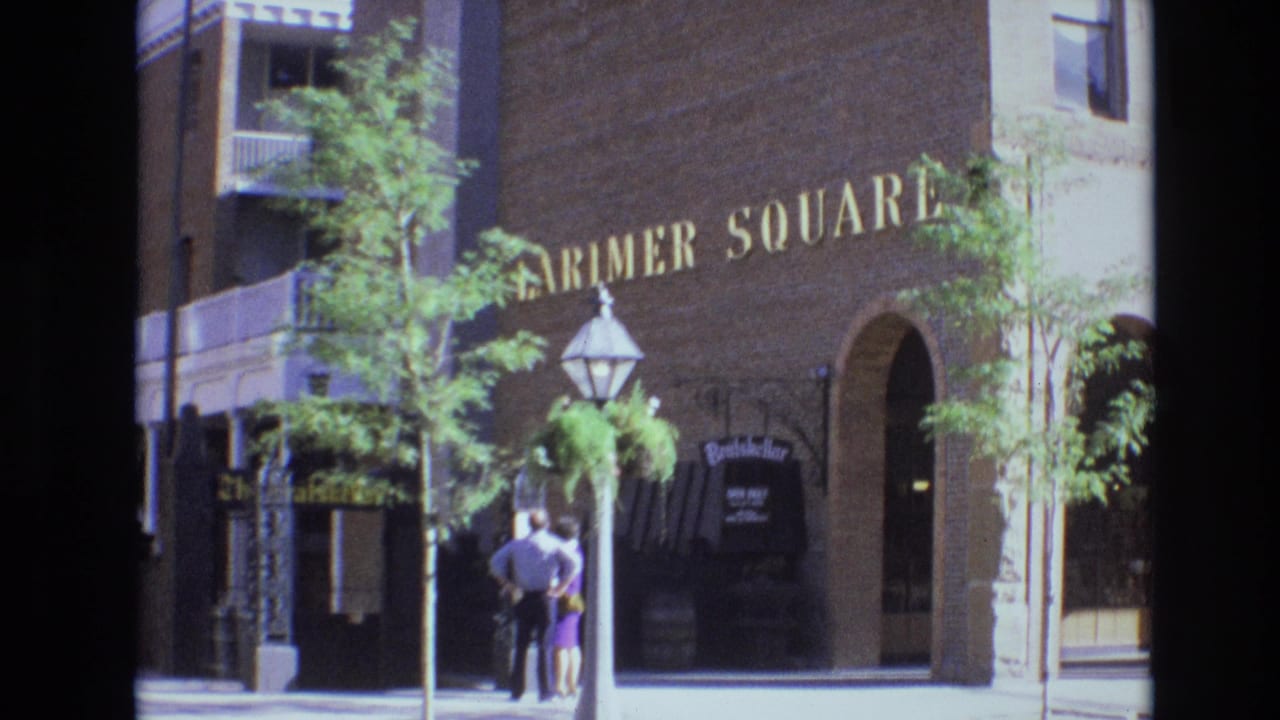 In supplied 1974 Denver footage, a man and woman stand near a lamppost and hanging plants outside a brick building with Farmer Square lettering.