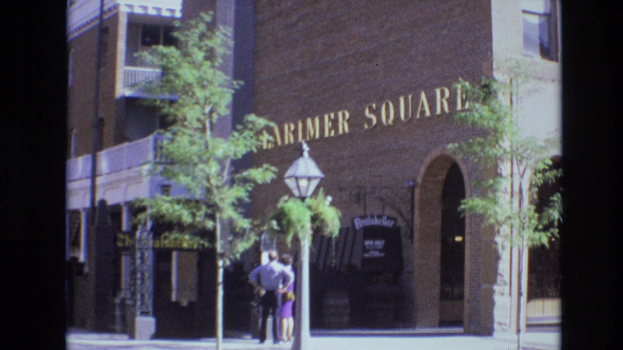 The frame shows the Farmer Square building facade in Denver, with a couple standing below gold lettering near a decorative streetlamp and shop awning.