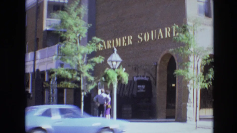 A light-colored car moves through the foreground while the couple remains on the sidewalk outside the brick Farmer Square facade.