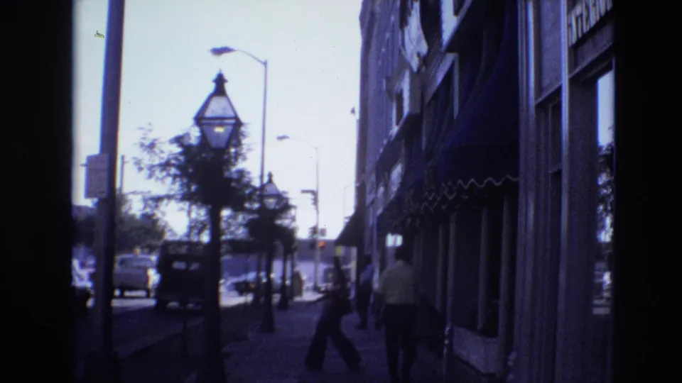 1974 Denver 8mm frame looking from shade along a sidewalk past storefront awnings, pedestrians, decorative street lamps, small trees, and period cars in bright street light.