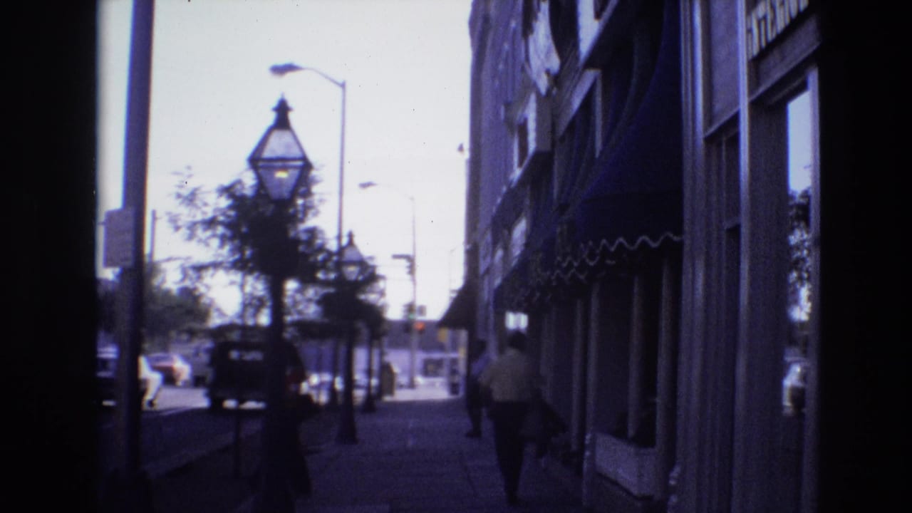 Sidewalk framed by dark edges with a pedestrian near storefronts and cars moving in the street beyond.