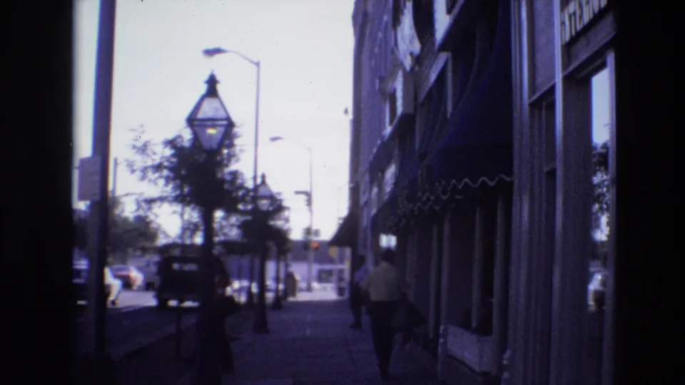 A 1974 Denver street scene filmed from a shaded sidewalk position, with a pedestrian near dark storefront awnings, vintage-style street lamps, trees, and cars in the bright roadway.