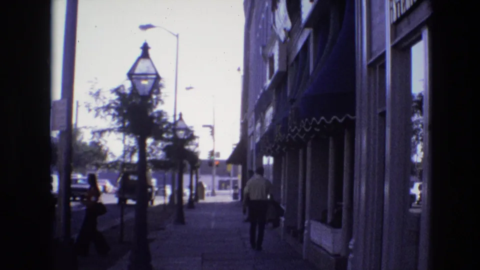 1974 Denver home-movie frame showing pedestrians on a shaded downtown sidewalk beside storefront windows and awnings, with decorative lamps, small trees, and cars in the sunlit street.
