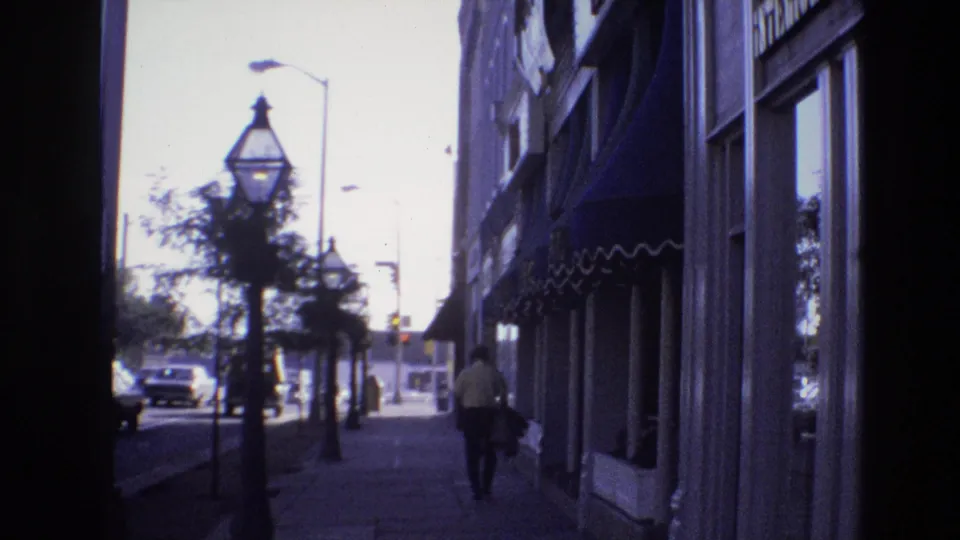Final 1974 Denver 8mm frame with a pedestrian walking away beside storefront awnings while decorative street lamps, trees, and period cars line the bright street beyond.