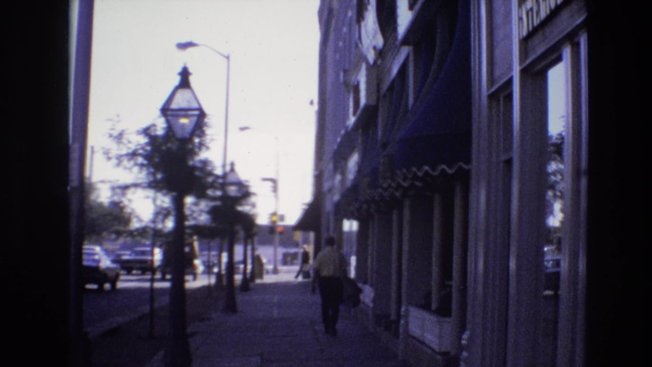 Doorway view onto a city sidewalk with storefronts, street lamps, cars, trees, and a pedestrian walking away.