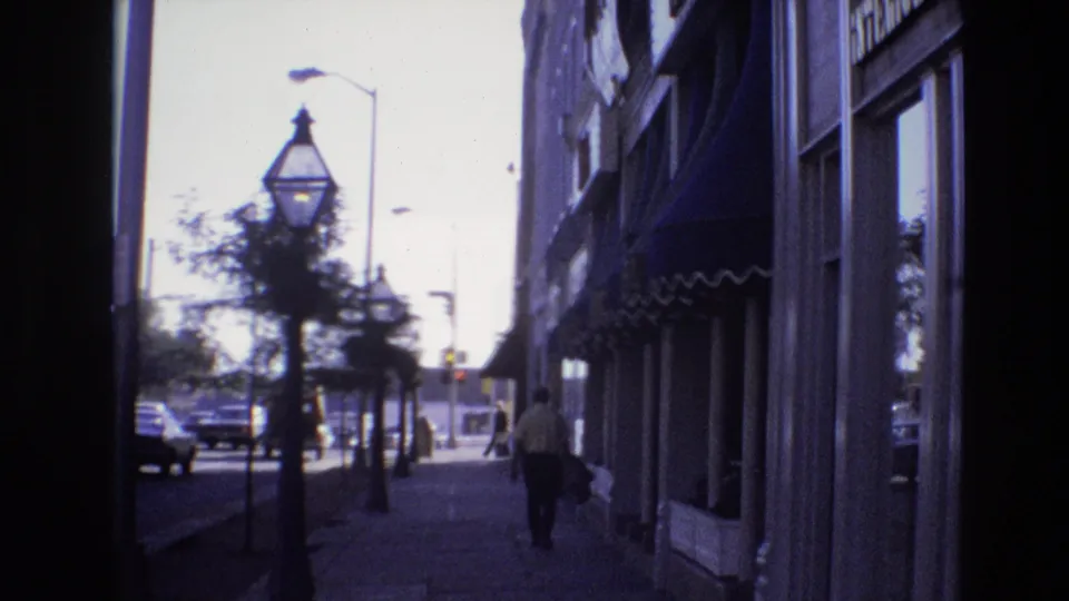 1974 Denver 8mm frame looking from a shaded doorway toward a sunlit sidewalk with storefronts, street lamps, passing cars, trees, and a pedestrian.
