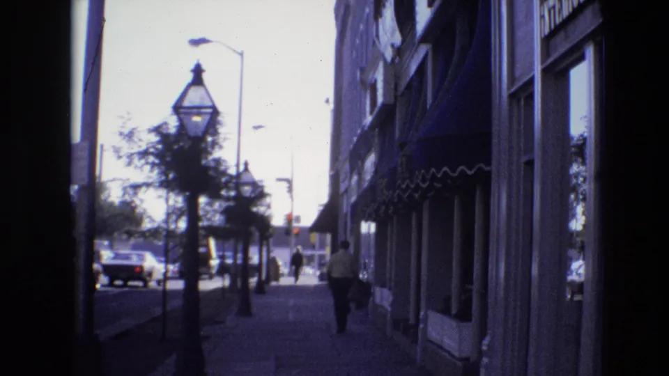 1974 Denver street view from a shaded entrance, with storefront awnings along the sidewalk, street lamps, trees, cars, and distant pedestrians.