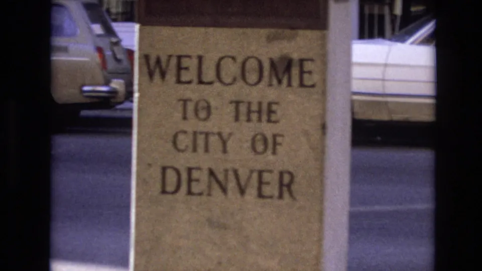 Close 1974 Denver 8mm frame of a stone welcome sign reading Welcome to the City of Denver, with vintage cars and a street behind it.