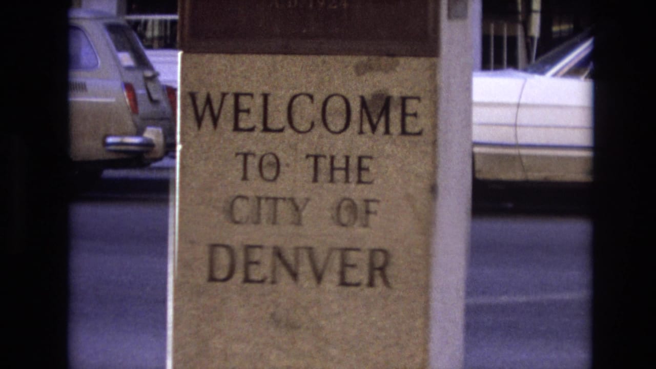 1974 Denver 8mm frame showing a close view of a stone WELCOME TO THE CITY OF DENVER sign with period cars visible across the street.