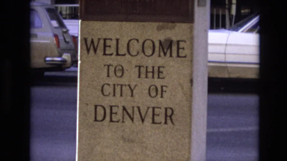 Close 8mm view of the Denver welcome sign, tightly framed on the lettering with parked 1970s cars and street pavement in the background.