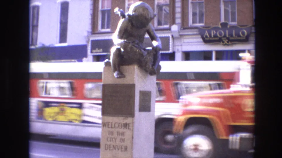 Wider 1974 Denver street frame showing the welcome sign below a bronze child statue, with a red fire truck, storefront windows, and an APOLLO sign in the background.
