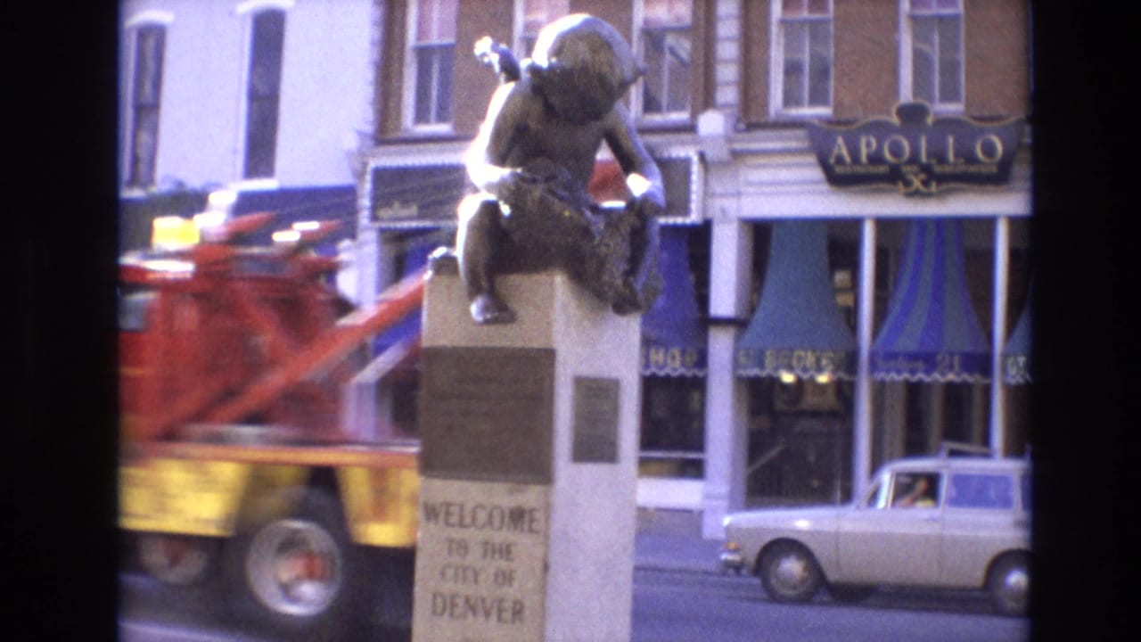 Denver welcome statue beside a street with storefronts and a passing vehicle.