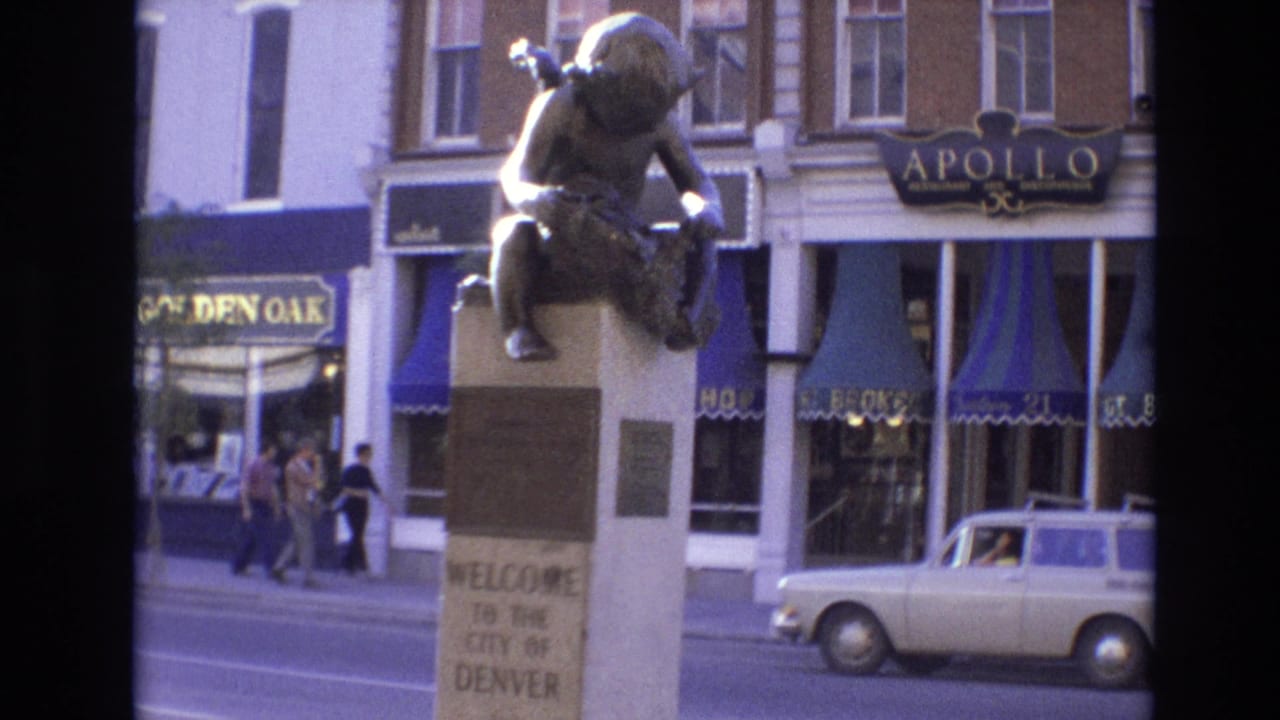 Welcome statue in front of shops, with pedestrians and a small car on the street.