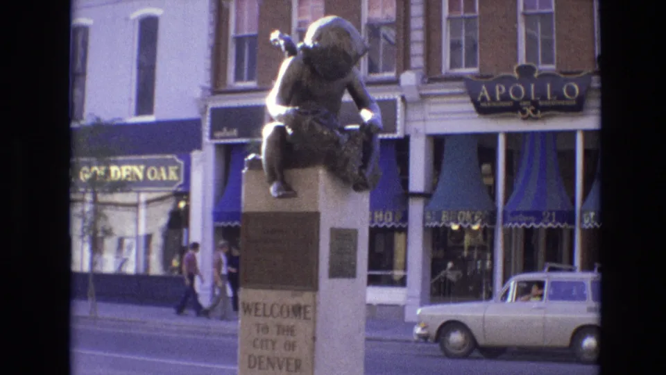 1974 Denver street scene with the welcome statue in the foreground, pedestrians along the sidewalk, blue-and-white awnings, shop windows, and a compact white car.