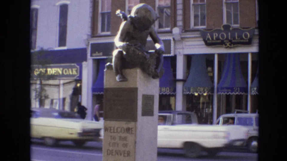 1974 Denver frame showing the welcome statue and commercial storefronts as a light-colored car moves through the foreground and a small white car remains near the curb.