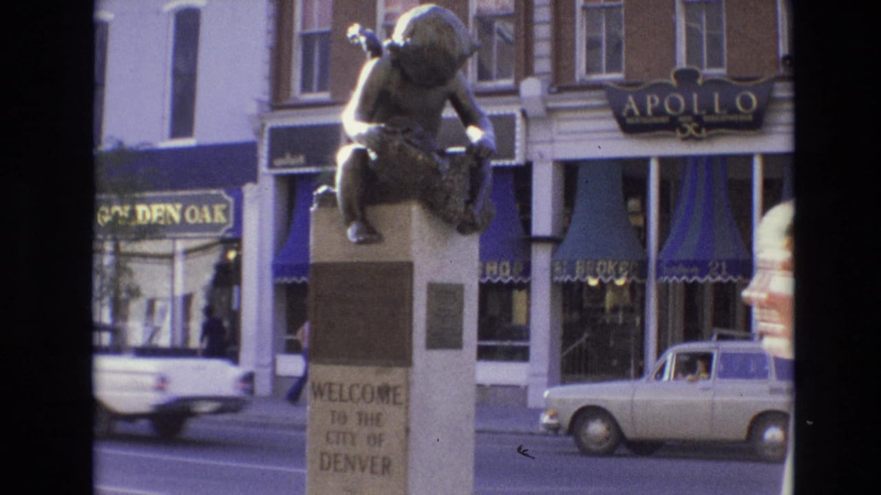 1974 Denver street view with a bronze child statue on a pedestal reading Welcome to the City of Denver, storefront signs, and period cars passing in the background.