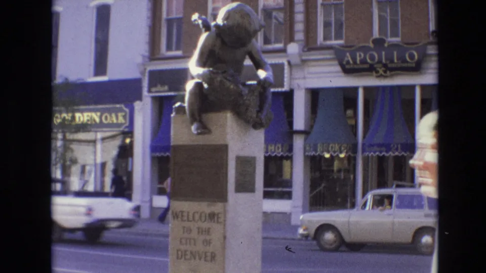 1974 Denver street view with a bronze child statue on a pedestal reading Welcome to the City of Denver, storefront signs, and period cars passing in the background.