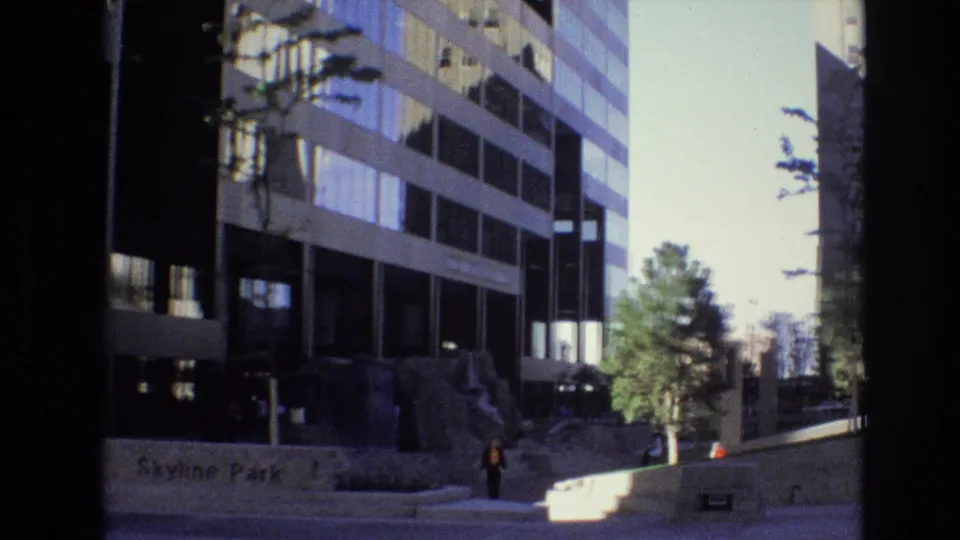 Denver office-building view identified by the wall text as Skyline Park, with a sidewalk, young trees, steps, and a pedestrian near the landscaped edge.