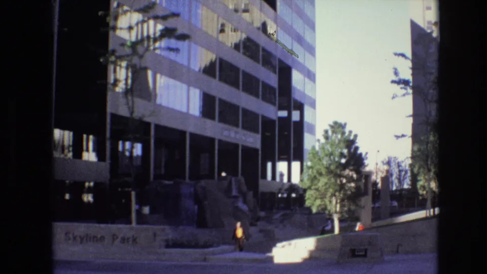1974 Denver view of the Skyline Park area, showing a modern office facade, wall lettering, a young tree, steps, and a pedestrian walking along the sidewalk.