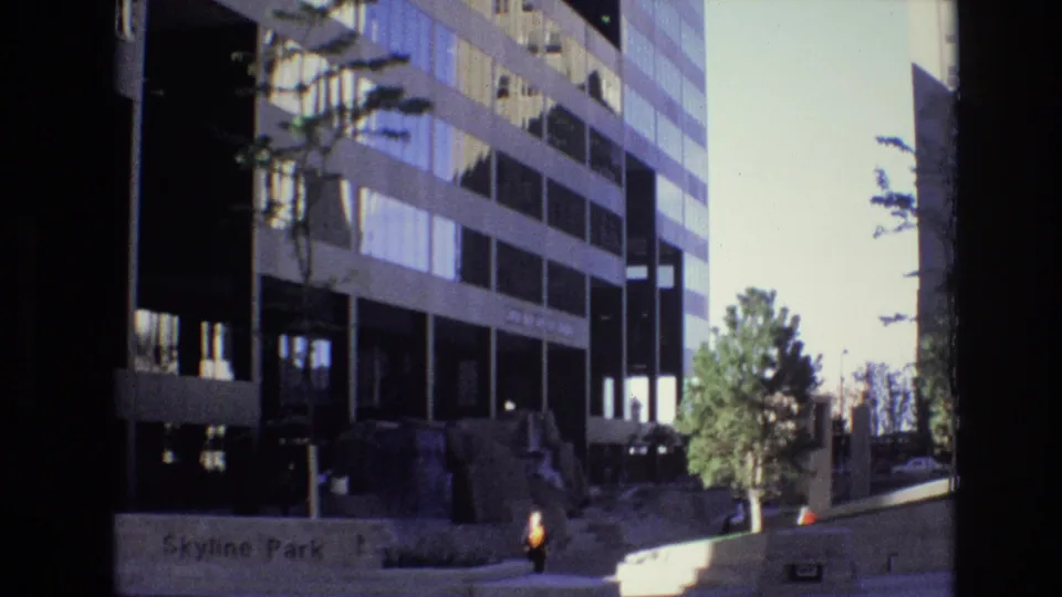 Denver Skyline Park streetscape with a multi-story office building, a pedestrian near the sidewalk, a landscaped tree, steps, and deep shadows from the surrounding buildings.