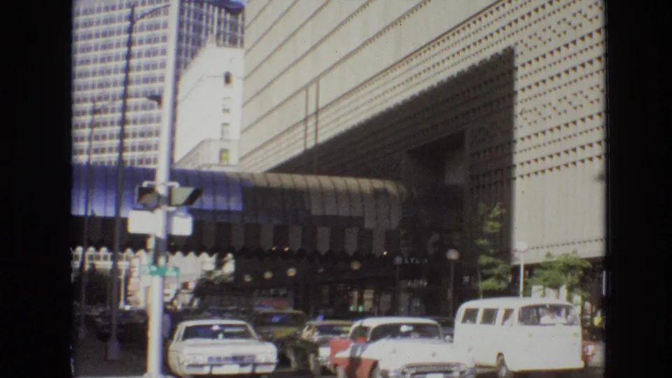 1974 Denver downtown traffic passes in front of a large modernist building, with pedestrians and street fixtures visible near the sidewalk.