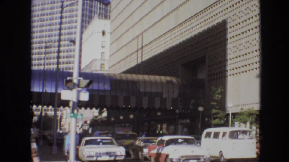 1974 Denver street scene with automobiles, pedestrians, and a patterned building facade viewed through soft 8mm home-movie framing.