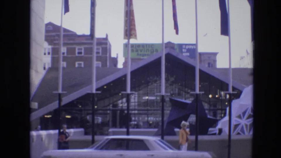 1974 Denver exterior view of a glass-fronted downtown entrance with flagpoles, signage, a parked car, and pedestrians in the foreground.