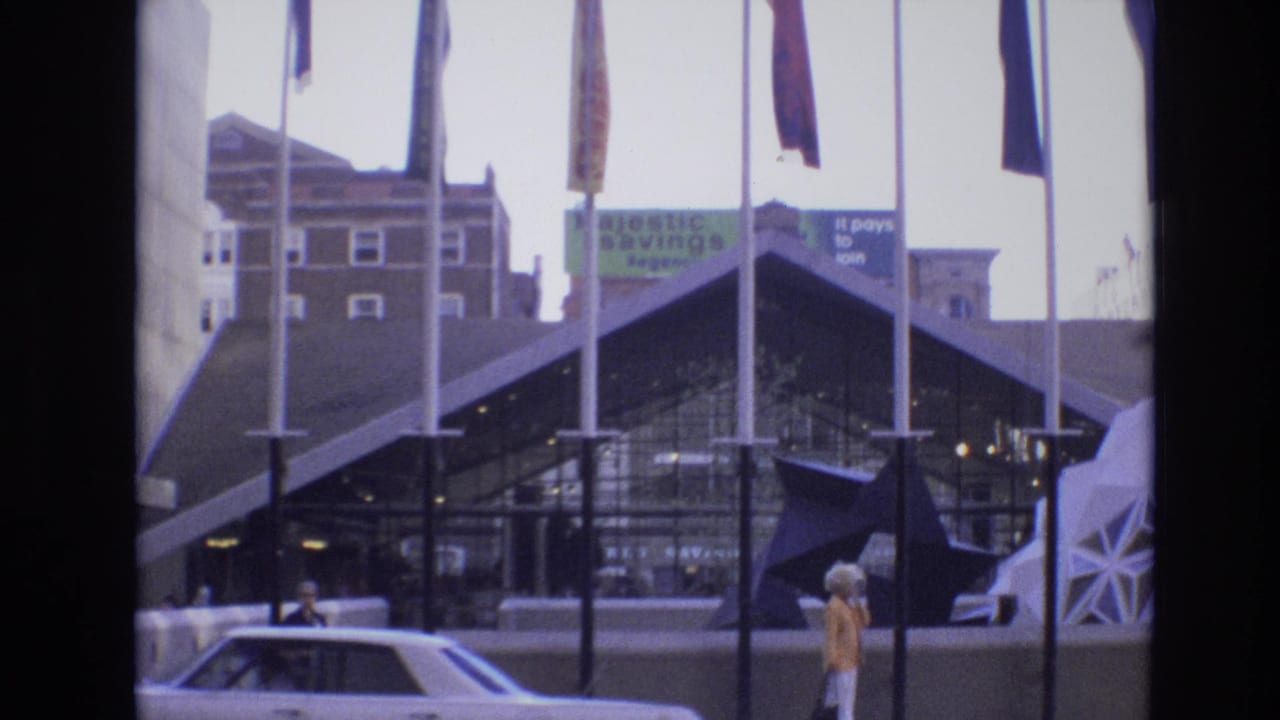 1974 Denver street view with tall international flagpoles, an angular glass-fronted building, background Majestic Savings signage, a parked car, and a woman walking past.